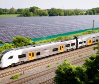Blick von schräg oben auf den Solarpark an der Bahntrasse mit Zug im Vordergrund